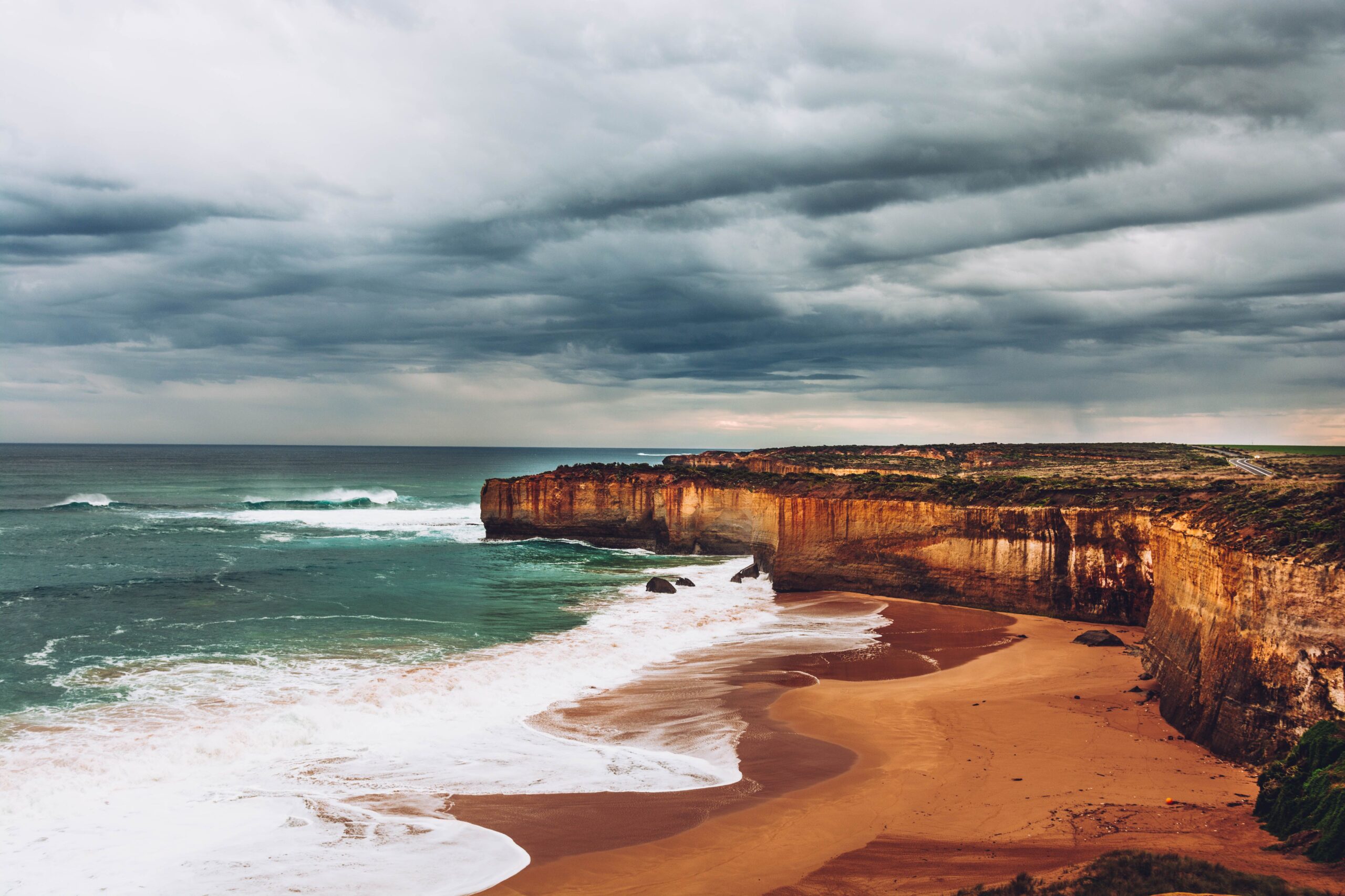 Dark storm clouds gather over the rugged limestone cliffs and sandy beach of Victoria's Great Ocean Road as waves roll in from the Southern Ocean. Rain falls offshore beneath the layered cloud deck, highlighting the dramatic coastal landscape and approaching weather.